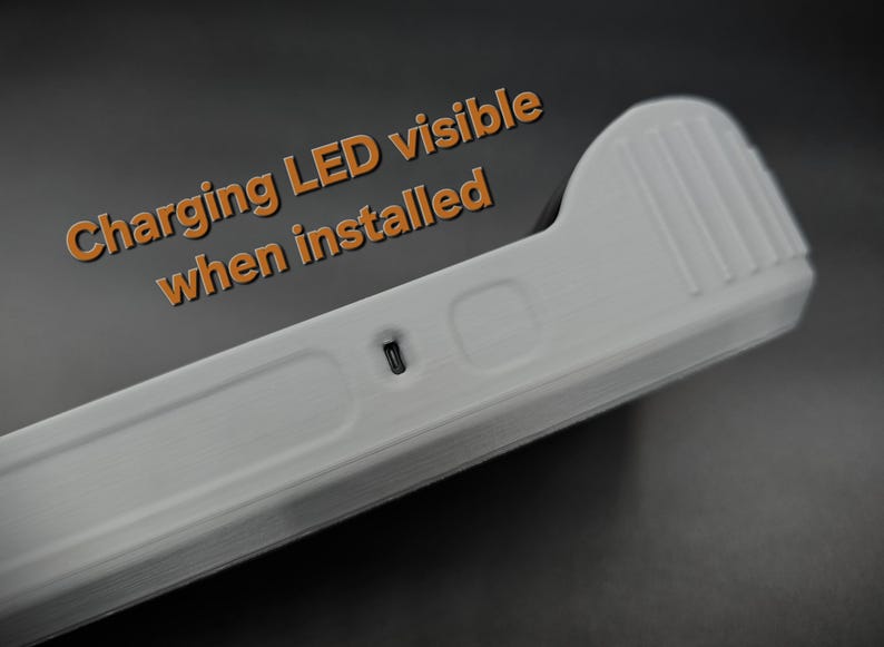 May include: A close-up of a white, rectangular device with a ridged top and a small, dark port. The text "Charging LED visible when installed" is in orange. The device appears to be made of plastic.