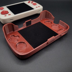 May include: A red plastic case for a handheld gaming device. The case has a textured surface and features a black screen protector, a button, and several circular cutouts.
