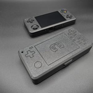 Puede incluir: Dos consolas de juegos portátiles de color gris oscuro se muestran sobre una superficie gris. La consola superior tiene una pantalla y botones. La consola inferior tiene una superficie texturizada con un diseño de mando.