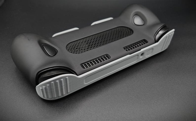 May include: A black and gray handheld gaming console case. The case has a textured black body with gray accents, including the grips and edges. It features a mesh vent and several button placements, designed for a comfortable grip and optimal gaming experience.
