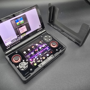 May include: A black handheld gaming console with a clamshell design, featuring two screens and multiple control buttons. The top screen displays a Nintendo DS game. A separate black stand is also visible.