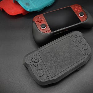 Puede incluir: Una consola de juegos portátil con carcasa roja translúcida y agarres negros, situada bajo una funda protectora negra texturizada. Dos carcasas de plástico adicionales en rojo y azul cerceta son visibles en la esquina superior izquierda.