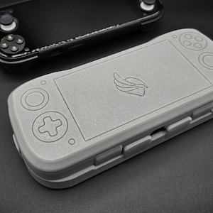 May include: A gray handheld gaming console case with a wing logo, featuring button outlines and a rectangular screen area. A black handheld gaming console is visible in the background. The case is designed to protect the console.
