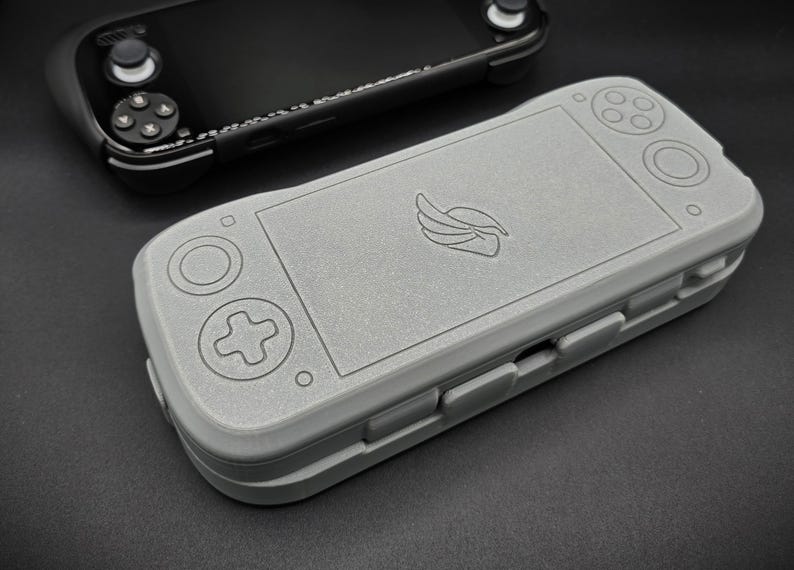 May include: A gray handheld gaming console case with a wing logo, featuring button outlines and a rectangular screen area. A black handheld gaming console is visible in the background. The case is designed to protect the console.