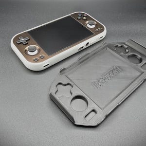 May include: A handheld gaming console with a white and gold-colored frame, black screen, and control buttons. A separate gray protective case is next to the console. The case has the text "RG477M" on it.