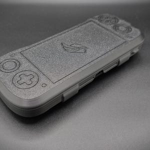 May include: A dark gray handheld gaming console with embossed buttons and a screen. The device features a directional pad, action buttons, and a logo on the screen. The console is made of a textured material.
