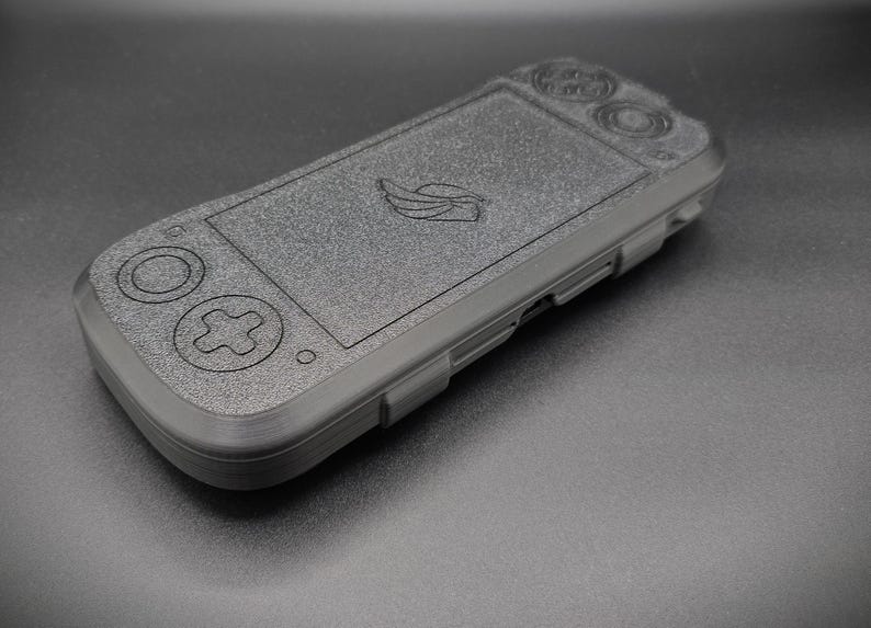 May include: A dark gray handheld gaming console with embossed buttons and a screen. The device features a directional pad, action buttons, and a logo on the screen. The console is made of a textured material.