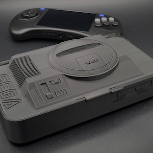 May include: A black Sega Genesis console replica with a top-loading CD drive and a Sega logo on the front. The console has two controller ports and a power switch.