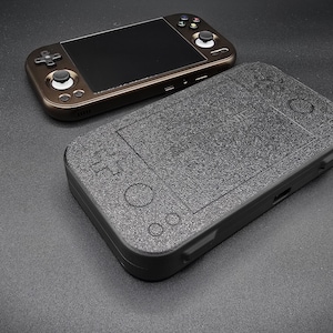 May include: Two handheld gaming consoles on a dark surface. One is brown with a black screen and buttons. The other is black with a textured surface and controller button outlines. The consoles are designed for portable gaming.