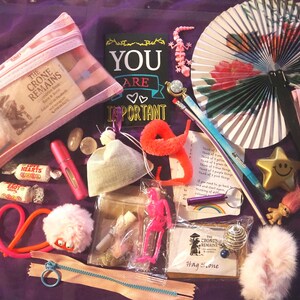 May include: A selection of small objects on a purple surface, including a clear zipped pouch, sweets, a fan, a star-shaped trinket, and a sign that reads "YOU ARE IMPORTANT". Other items include a pink perfume bottle, a pink lizard, and a fluffy ball.