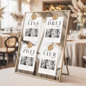 Puede incluir: Un expositor con marco dorado con cuatro tarjetas, cada una sujeta por un sello de madera. Las tarjetas muestran los números "EINS", "DREI", "ZWEI" y "VIER" en texto negro, junto con fotos en blanco y negro de un niño. El expositor está sobre una superficie blanca.