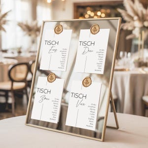 Puede incluir: Un plano de asientos con marco dorado con cuatro tarjetas blancas, cada una etiquetada como "TISCH" y un número. Las tarjetas se mantienen en su lugar mediante sellos de cera dorados. Cada tarjeta enumera nombres debajo del número de la mesa. El fondo es un entorno de evento borroso.