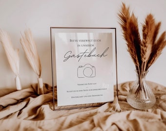 May include: A white sign with a gold frame, with the text "Bitte verewigt euch in unserem Gästebuch" and a line drawing of a camera. The text below the camera says "Nehmt ein Foto auf sucht euch eine Seite zum einkleben raus hinterlasst uns eine Erinnerung."