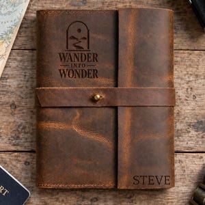 May include: A brown leather journal featuring a horizontal strap with a metal stud closure. The cover is engraved with the text "WANDER INTO WONDER" above an arch icon and the name "STEVE" in the bottom right corner, showcasing a rustic texture.