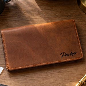 May include: A rectangular brown leather checkbook cover featuring the name Parker embossed in a dark script font on the front. The cover is positioned on a wooden surface alongside a gold pen and an open check register with visible grid lines.