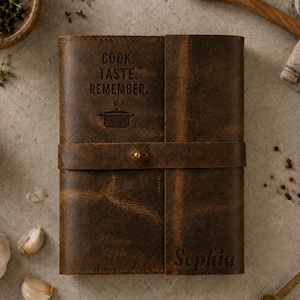 May include: A dark brown leather journal with a horizontal strap and a brass button closure. The cover is embossed with the text "COOK. TASTE. REMEMBER." above a cooking pot icon, and the name "Sophia" is embossed in the bottom right corner.