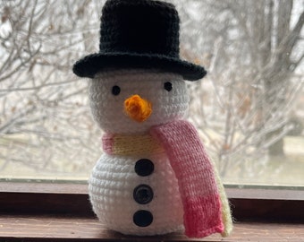 May include: A crocheted snowman wearing a black top hat and a pink and yellow scarf. The snowman has black eyes and a bright orange nose.