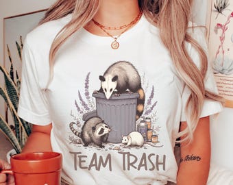 May include: White t-shirt with a graphic of a possum, a raccoon, and a rat standing around a trash can. The text "Team Trash" is printed below the graphic.