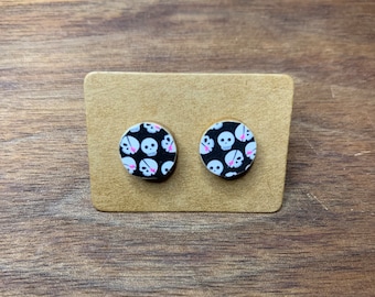 May include: A pair of round stud earrings with a black background and a white skull pattern with pink accents.