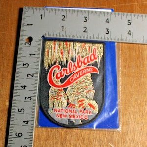 Puede incluir: Parche bordado con las palabras "Carlsbad Caverns" en escritura roja, y una representación de formaciones de cuevas. El parche es negro con detalles dorados y rojos, y el texto "National Park New Mexico" está debajo. El parche mide aproximadamente 7.6 cm de largo.