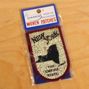 May include: Gold iron-on patch with a black outline of the state of New York. The patch says "New York" and "The Empire State" in black text.
