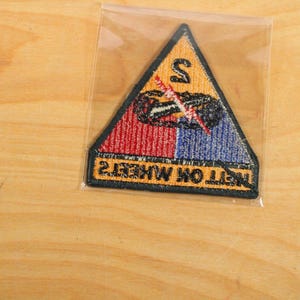 May include: A triangular patch with a black border, featuring a red and blue vertical split design. The patch has the text "NETT OK WHEELS" and a graphic of a vehicle with a lightning bolt. The top section is yellow with a black "S".