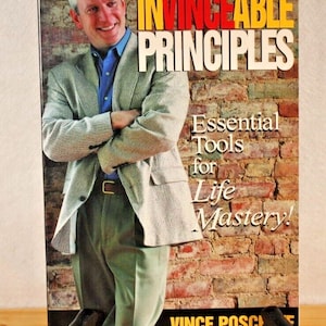 May include: A book titled "Invincible Principles: Essential Tools for Life Mastery!" by Vince Poscente. The cover features a man in a light gray blazer with a blue shirt, standing in front of a brick wall.