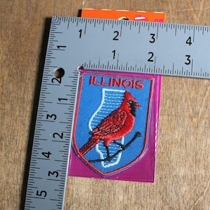 Op de afbeelding: Geborduurde patch met een rode kardinaal op een tak, tegen een blauwe achtergrond met de omtrek van Illinois en het woord "ILLINOIS". De patch is ongeveer 5 cm breed.