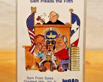 以下が含まれることがあります： 「Sam Pleads the Fifth」と題された、漫画のイラストが描かれたヴィンテージカセットテープ。テープには「Sam From Sales Greatest Hits, Vol. V」の文字と、BASFとWBAP Radio 820のロゴが描かれています。カセットは黒いスタンドで支えられています。