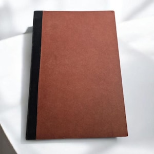 May include: A closed, rectangular notebook with a reddish-brown cover and a black spine. The notebook is lying on a white surface, with the light creating shadows on the cover.
