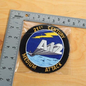 Puede incluir: Un parche circular con el texto "21st Century Medium Attack A12" en blanco y amarillo. El parche presenta un fondo azul con un diseño estilizado de avión y rayo. Una regla metálica está al lado del parche.