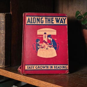 Pode incluir: Um livro vintage intitulado "Along the Way" com capa vermelha. A capa apresenta uma ilustração azul e branca de uma pessoa lendo para uma criança em uma mesa. O texto "Easy Growth in Reading" está na parte inferior.