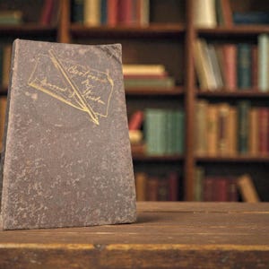 May include: A vintage book with a worn, textured cover. The cover is a muted gray-brown color, with gold lettering and a pen illustration. The title reads "Cartoons by Sargent J. Preston." The book is positioned on a wooden surface, with a blurred bookshelf in the background.