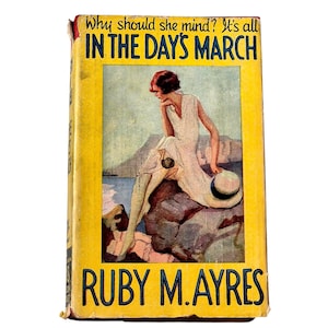 May include: A vintage book titled In The Days March by Ruby M. Ayres with a yellow cover. The front features an illustration of a person in a white dress sitting on a rock by the sea, holding a straw hat. The text reads Why should she mind? It's all IN THE DAY'S MARCH and RUBY M. AYRES.