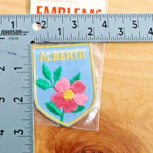 May include: A light blue embroidered patch featuring the word "ALBERTA" in yellow above a pink and yellow flower with green leaves. The patch is approximately 2 inches wide and 2.5 inches tall.