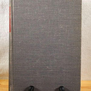 Może przedstawiać: Książka w twardej szarej oprawie z teksturowaną okładką, prezentowana na czarnym stojaku. Grzbiet książki ma czerwony akcent. Książka stoi pionowo, prezentując swoją okładkę.