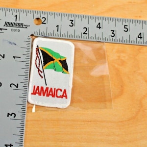 May include: A rectangular patch featuring the Jamaican flag, with a black and yellow diagonal cross on a green background. The word "JAMAICA" is printed in red below the flag. The patch is approximately 2 inches long.