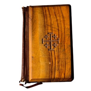 May include: A rectangular book with a light brown wood cover featuring a dark brown leather spine. The front cover displays a centered, embossed Jerusalem cross symbol. The wood grain is visible, and a thin brown ribbon bookmark extends from the bottom.