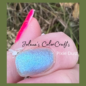 May include: A fingernail with a light blue nail polish with a glittery finish. The nail polish is labeled "Jolene's Color Crafts Pixie Dust".