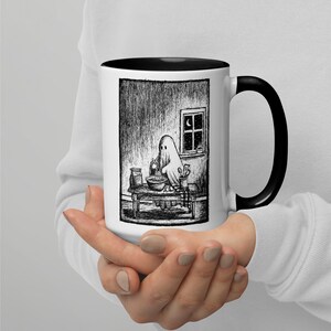 May include: A white ceramic mug with a black handle and interior rim, featuring a detailed black and white ink-style illustration of a ghost wearing a sheet while baking at a wooden table in a room with a window showing a crescent moon.