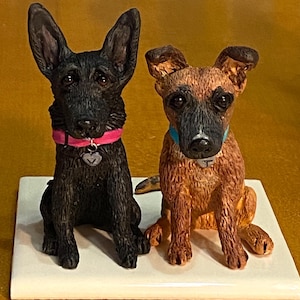 May include: Two ceramic dog figurines, one black and one brown, sitting side-by-side on a white base. Both dogs have collars and tags.