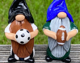 Puede incluir: Dos gnomos, uno con un sombrero negro y sosteniendo una pelota de fútbol, el otro con un sombrero azul y sosteniendo una pelota de fútbol americano. Ambos gnomos llevan camisas a rayas blancas y negras y pantalones marrones.