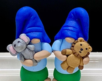 Puede incluir: Dos figuras de gnomos azules y blancos sosteniendo animales de peluche. El gnomo de la izquierda sostiene un elefante gris y el gnomo de la derecha sostiene un león marrón.