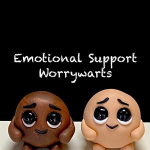 May include: Two small, round, clay figures with big eyes and smiling mouths. The figures are brown and beige and are holding their hands in front of them. The text "Emotional Support Worrywarts" is written above the figures.