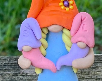 Puede incluir: Una caprichosa figura de gnomo hecha de arcilla. El gnomo lleva un sombrero naranja con una flor amarilla y centro morado, un vestido azul y zapatos naranjas. Sostiene dos pequeños sombreros, uno lavanda con una "S" y otro rosa con una "L".