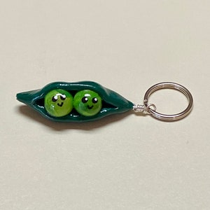 May include: A green pea pod keychain with two smiling peas inside. The keychain is made of polymer clay and has a silver metal ring.