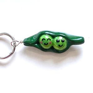 May include: A handmade keychain featuring a green pea pod with two smiling peas. The peas have black eyes and rosy cheeks. The keychain has a silver-colored metal ring.