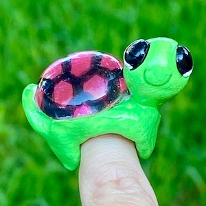 May include: A green turtle ring with a red and black shell design. The turtle's head has large black eyes and a smiling expression. The ring is worn on a finger, showcasing the handcrafted jewelry.