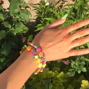 May include: A close-up of a person's wrist wearing three colourful bracelets made of fruit-shaped beads. The bracelets are stacked on top of each other and feature a variety of fruits, including strawberries, lemons, oranges, and pineapples. The person's nails are painted a light green colour.