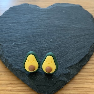 Può includere: Due orecchini a forma di avocado con un viso sorridente e un seme marrone. Gli orecchini sono verdi e gialli e sono su una superficie nera a forma di cuore.